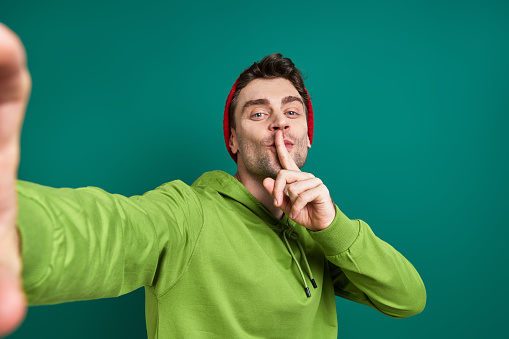 Handsome man making selfie and holding finger on lips while standing against green background