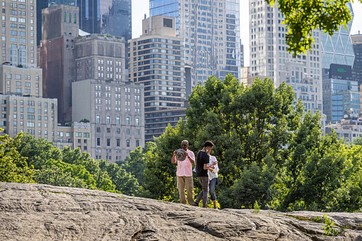 Central Park, Manhattan, New York, NY, USA - June 29, 2022: Three people standing on a rock in front of a tree and the high rise skyscrapers