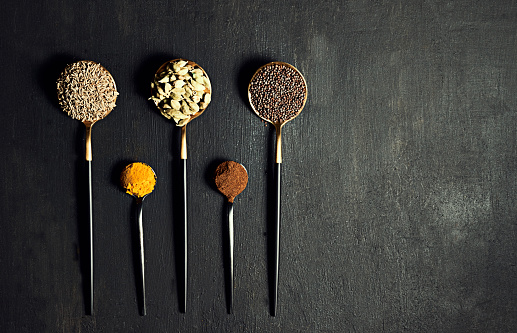 Above view of spices, seasoning and ingredients on flat lay spoons against black copy space background. Detail of mustard or fennel seeds, turmeric and cardamoms for cooking tasty indian curry meals