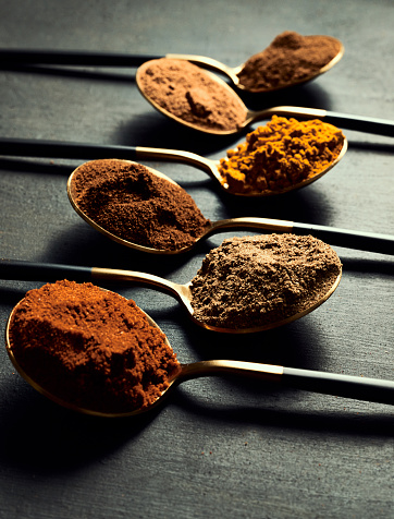 Spices, flavors and curry powder lined up in spoons on a black table for cooking a tasty meal. Ingredients for a flavorful dish on utensils with a rustic background. An assortment of Indian seasoning