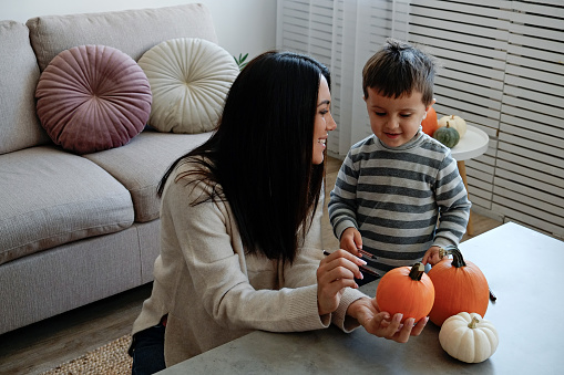 Young beautiful mother with her three year old son preparing decorations for halloween at home. Woman spending quality time with her child, carving pumpkins. Close up, copy space, background.