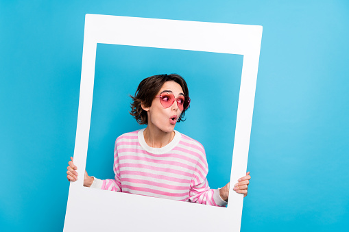 Photo of impressed astonished lady look empty space hold white picture frame isolated on blue color background.