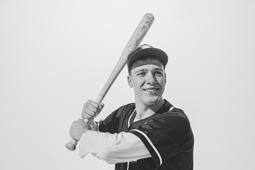 Black and white portrait of smiling young man, baseball player in uniform with bat preparing to hit ball. Motivated sportsman. Concept of sport, retro style, 20s, fashion, action, college sport, youth