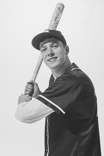 Black and white portrait of smiling young man, baseball player in uniform with bat preparing to hit ball. Student lifestyle. Concept of sport, retro style, 20s, fashion, action, college sport, youth