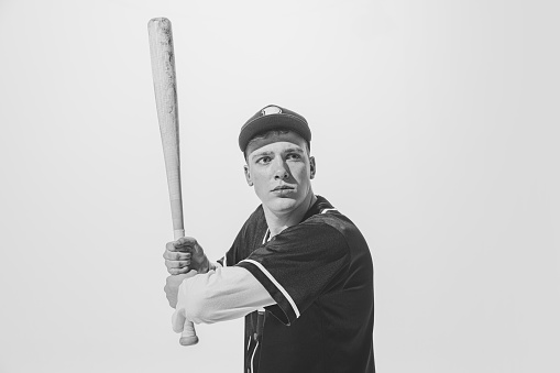 Black and white portrait of concentrated young man, baseball player in uniform with bat preparing to hit ball. Concept of sport, retro style, 20s, fashion, action, college sport, youth culture