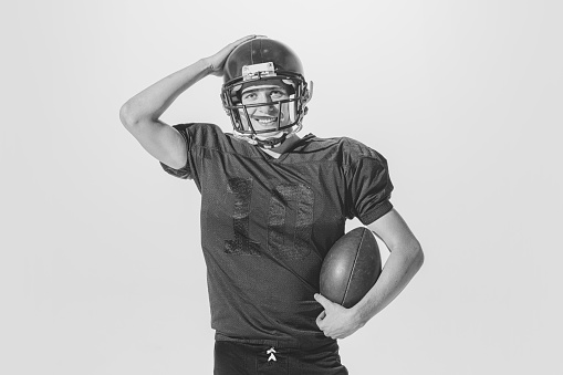 Portrait of young smiling man, american football player posing in uniform and helmet with ball. Black and white photography. Concept of sport, retro style, 20s, fashion, action, college sport, youth
