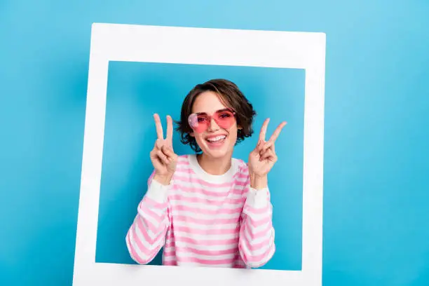 Photo of attractive cheerful female in heart spectacles show v-sign through white frame cadre isolated on blue color background.