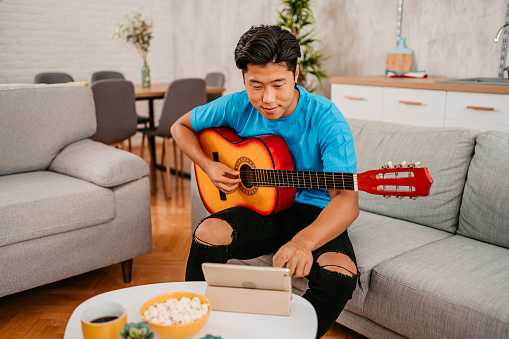 Handsome young Chinese man watching a tutorial on how to play an acoustic guitar, on tablet, in the living room.