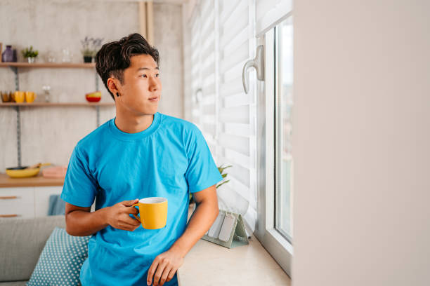Handsome young Chinese man drinking coffee and looking out the window of his apartment.