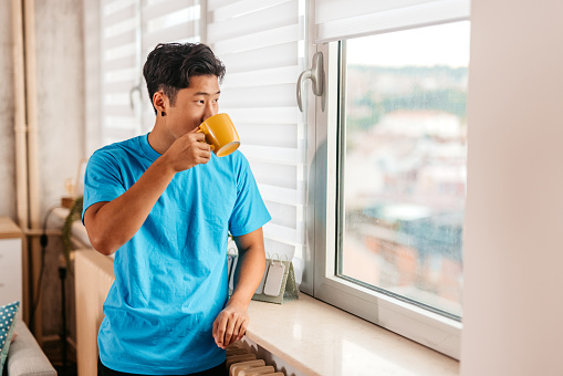 Handsome young Chinese man drinking coffee and looking out the window of his apartment.