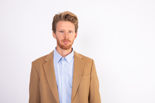 Portrait of happy young Caucasian businessman looking at camera. Bearded man wearing blue shirt and beige woolen suit coat standing and smiling. Successful businessman concept