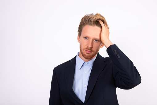 Portrait of stressed young businessman clutching head. Bearded Caucasian man wearing blue shirt and black suit coat looking at camera with troubled or tired expression. Problem and stress concept