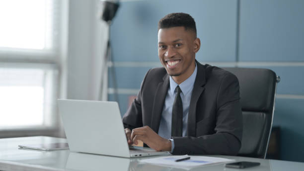 istock African Businessman Smiling at Camera while using Laptop in Office 1413824609