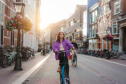 Young Caucasian woman in purple coat riding bicycle in the city centre  at sunset