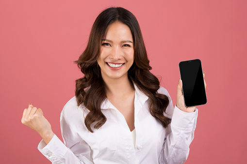 Asian woman wearing a white shirt a Very excited, very happy  showing a blank cell phone screen while standing split over pink background.