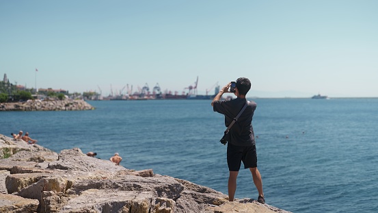 A young male tourist is taking photos and videos with his mobile phone by the sea.