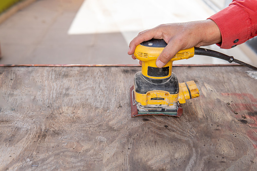 Close-up of a man's hand sanding a piece of wood with an electric sander on a patio outdoors