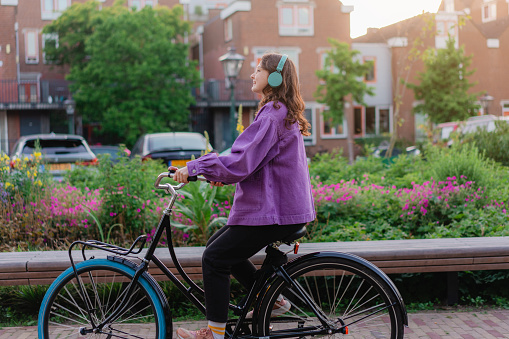 Young Caucasian woman riding bicycle in the city at sunset while listening to music in headphones