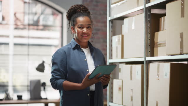 Portrait of a Successful Employee Checking Inventory, Writing in Tablet Computer. Black Woman Posing for Camera and Smiling in a Warehouse Storeroom with Orders Ready for Shipment.