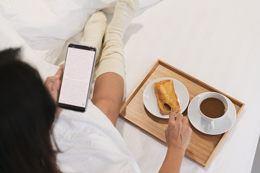 Young Asian woman relax on bed while using mobile phone and eat breakfast. Technology, lifestyle, food and beverage concept. Top view and copy space.