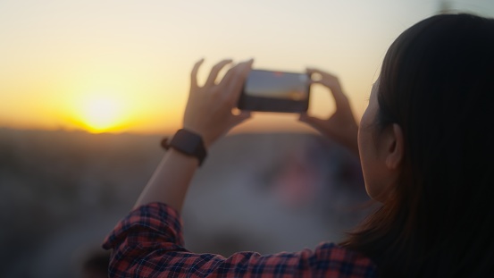 A young female traveler is taking photos and videos of sunset with her mobile phone on a hill in a historical old town.