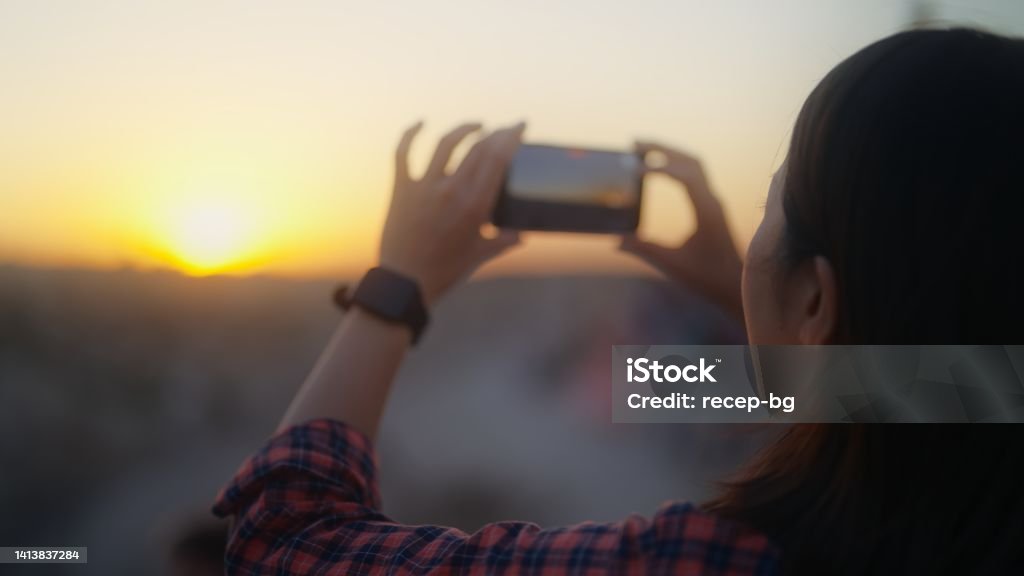 Young female traveler taking photos of sunset with her mobile phone on hill in historical old town A young female traveler is taking photos and videos of sunset with her mobile phone on a hill in a historical old town. Hiking Stock Photo