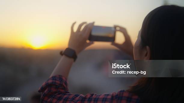 Young Female Traveler Taking Photos Of Sunset With Her Mobile Phone On Hill In Historical Old Town Stock Photo - Download Image Now