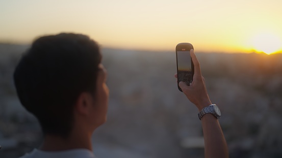 A young male tourist is taking photos and videos with his mobile phone on top of a hill in a historical old town.