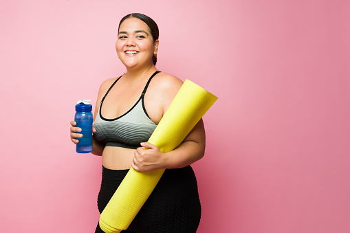 Attractive obese woman laughing after finishing her fitness workout and drinking from a water bottle against a pink background with copy space