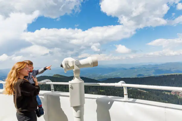 Photo of Mother shows son landscapes in valley of Rhodope Mountains and sky through telescope on observation tower of Snezhan