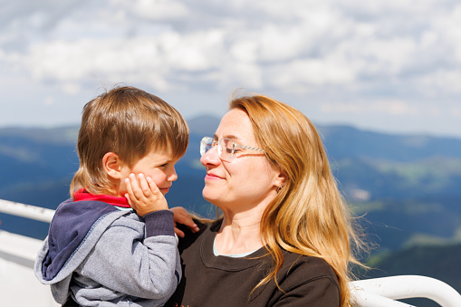 Caring mother hugs and loves her little son-toddler on high observation tower on top of Snezhan, against backdrop of mountain forest valley of Rhodope Mountains and bright blue cloudy sky