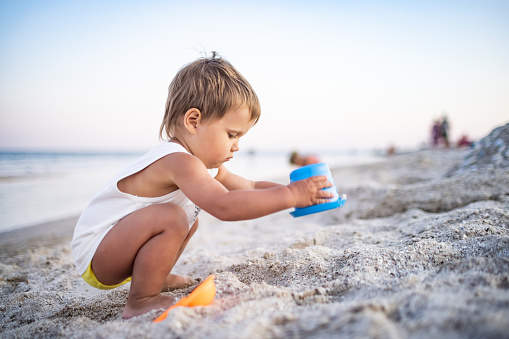 A little cheerful, enthusiastic boy plays with his toys on a sea sandy beach, building beads and turrets, smiling at someone behind the scenes on a bright sunny summer vacation