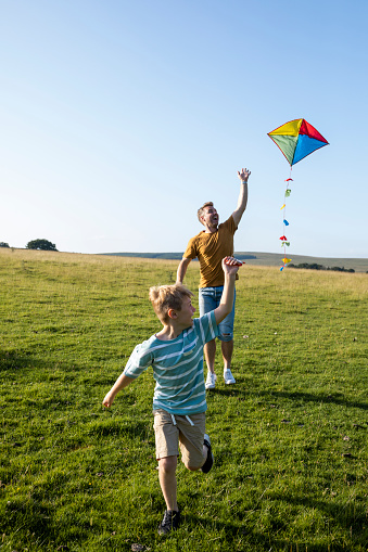 A dad and his young son having a great time and running on the grass in a field in the North East of England. The young boy is pulling a multicoloured kite and smiling with glee as they enjoy quality time together.