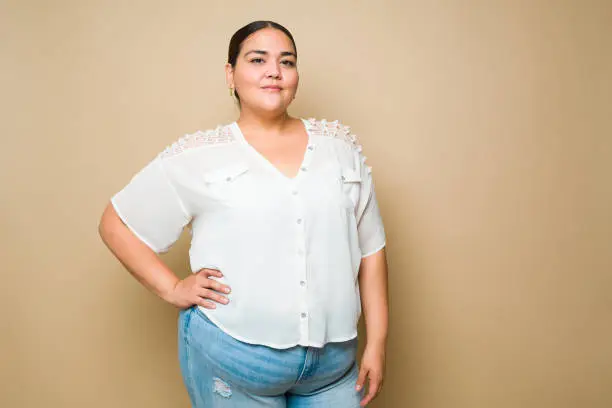 Attractive hispanic obese woman wearing casual clothes with a hand on her hip and smiling against a yellow background