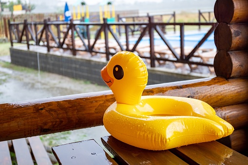 A yellow inflatable duck, lies on massive wooden table in cozy gazebo of one of houses at recreation center against the backdrop of a large pool covered with heavy pouring rain