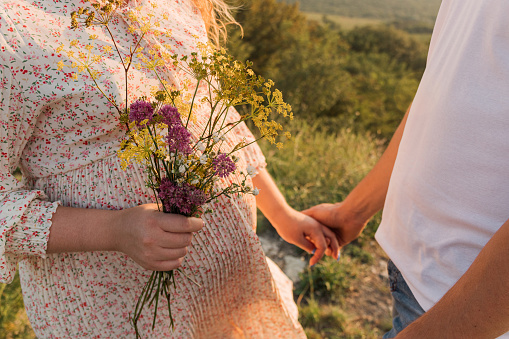 Pregnant woman holds wildflowers in her hand, holding man's hand with other hand. Selective focus. Picture for articles about pregnancy, care, family, psychology.