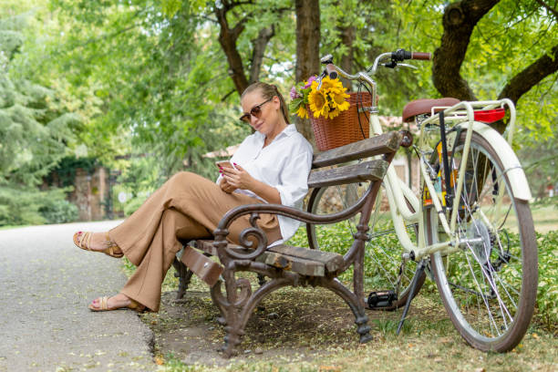Young woman texting on smartphone and sitting on a bench on a sunny day, beside the bike with flower basket.