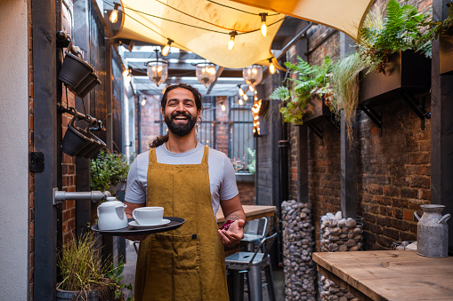 Portrait of a waiter looking at the camera smiling while holding a tray with a teapot and cup with saucer on it. He is wearing an apron and is working outdoors in a pub garden in the North East of England.