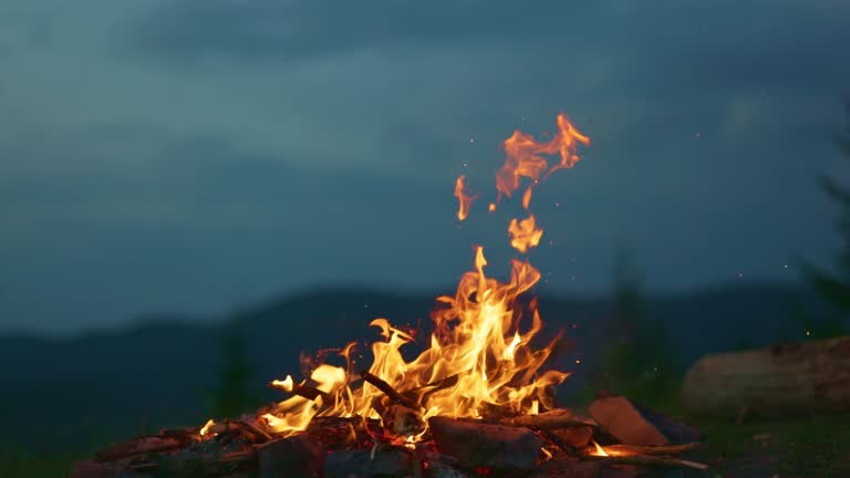 Campfire at dusk against the backdrop of mountains and forest. Tourist bonfire with sparks flying in the air, slow motion shot