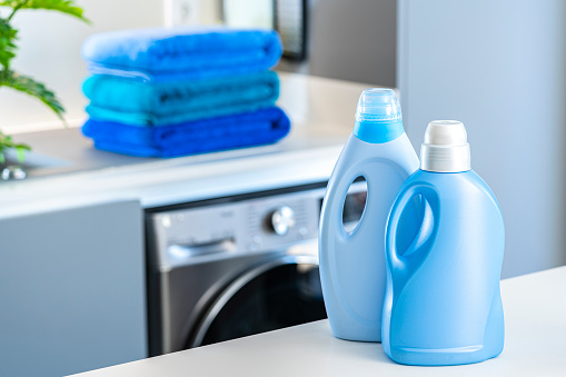 Blue fabric softener and detergent bottles shot on kitchen counter with washing machine and blue towels at background. Selective focus on bottles. Digital capture taken with Sony A7rII and Sony FE 90mm  digital capture taken with Sony A7rII and Sony FE 90mm f2.8 macro G OSS lens