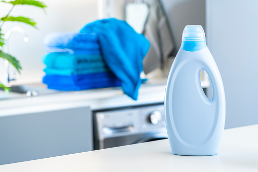 Blue fabric softener or detergent bottle shot on kitchen counter with washing machine and blue towels at background. Selective focus on bottle. Digital capture taken with Sony A7rII and Sony FE 90mm  digital capture taken with Sony A7rII and Sony FE 90mm f2.8 macro G OSS lens