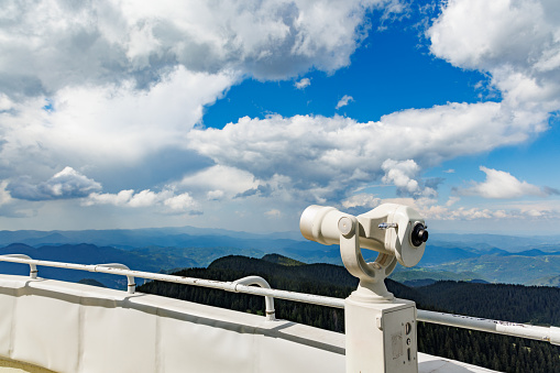 A modern metal orbital telescope of white color on a high observation tourist tower above a mountain forest high valley of the Rhodope mountains, against a sunny blue cloudy sky