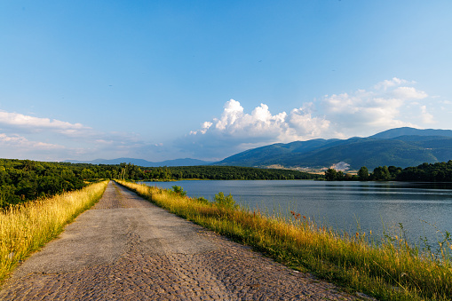 Even long vegetative road of sand and clay passes through swampy blue mountain lake, against backdrop of mountain valley of Rhodope Mountains with dense dark spruce forests and sunny cloudy sky