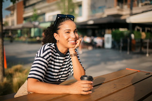 Happy young adult woman sitting in an outdoor coffee shop, enjoying a cup of coffee and relaxing