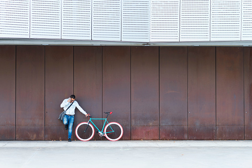 Young male with sunglasses and shoulder bag standing next to a fixed gear bike while calling by phone