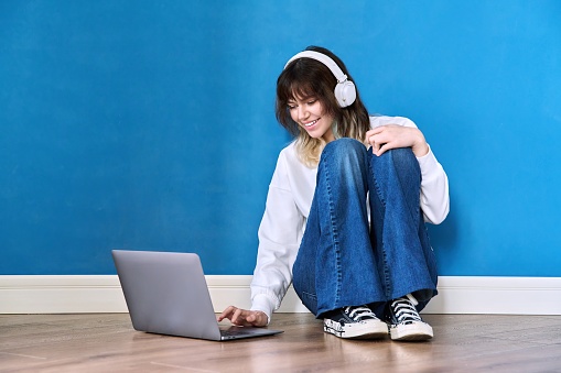 Beautiful teenage female in headphones sitting on floor with laptop on blue background. Hipster teenager looking at laptop screen. Youth, lifestyle, leisure, education, technology concept