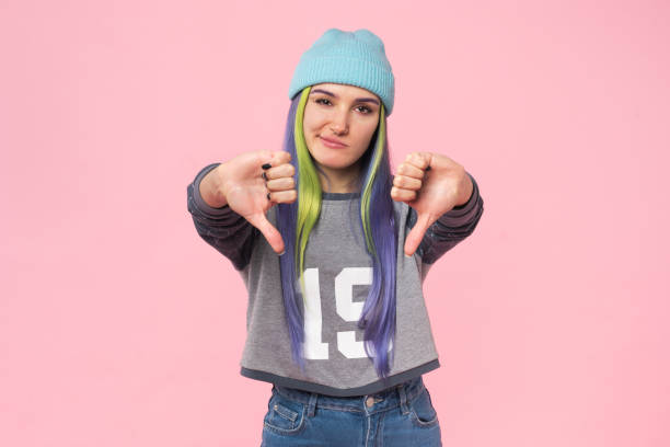Disappointed sad angry offended caucasian young teenage woman girl hipster showing thumbs down dislike isolated in pink background stock photo