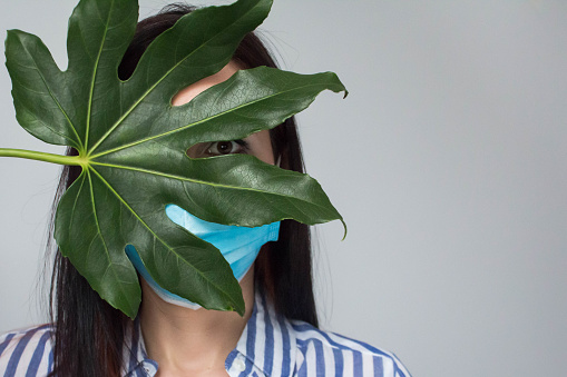 Beautiful woman hides face in a medical mask and open her eyes with big green tropical leaf, Faceless