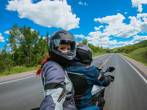 Motorcycle passenger young woman in a helmet makes selfie on action camera while riding on back of motorcycle on an empty landscape road.