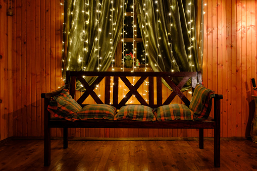 Glowing yellow festive LED garland hanging on the curtains. Bench with pillows against the background of a window with curtains and LED garland.